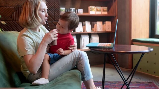 Tired mother working oh her laptop, holding her toddler son and drinking coffee sitting in a modern coffee shop. The problem of combining motherhood and work
