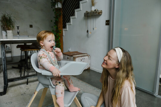 Mother Smiling While Feeding Her Baby Sitting in a High Chair