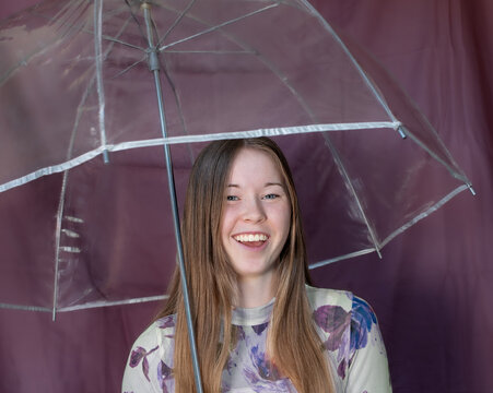 Close-up of One Smiling Young Woman Holding Clear Umbrella, Egg Plant Colored Background, squarish