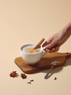 Hand Stirring Cinnamon Stick in Cup of Spiced Latte on Wooden Board