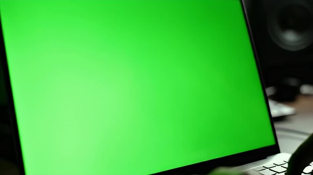 A person types on laptop with green screen. Dark room, hands focused. Speaker in background