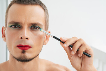 Fototapeta premium adult caucasian gay man with makeup and cosmetics on face. Transgender male with red lipstick applied eye shadow sparkle on his eyes with a brush on a white background