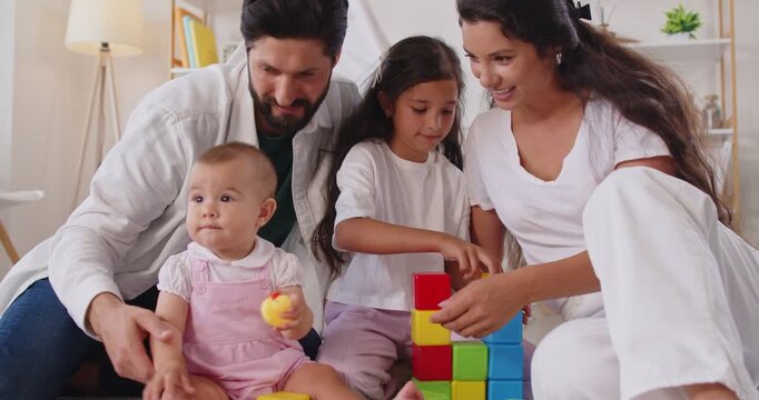 Happy family playing with block toys at home to build tower and develop motor skills of children. Playful baby toddler and girl play learning game with support of mother and father, sitting on floor