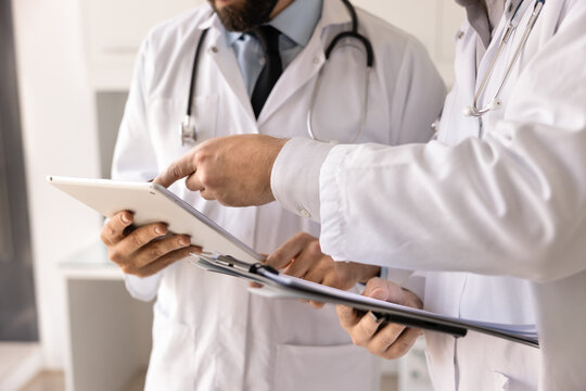 Digital and paper tools in medicine. Close up two male doctors medical workers in white coats discuss patient information analyze files on electronic tablet review written case history on clipboard