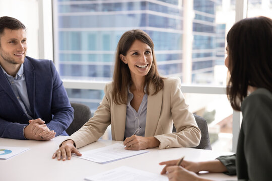 Professional middle-aged woman in formal attire sits at conference table with business partners, exude confidence, holds pen, ready to sign contract or agreement paper. Merger, commercial deal making