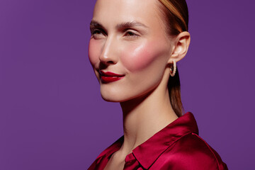 Portrait of confident woman with red lipstick and shiny skin wearing red blouse and gold hoop earrings on purple background. Concept of beauty, fashion, and elegance. © SHOTPRIME STUDIO