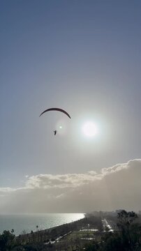 Paraglider soaring in the sky towards bright afternoon sun. Stunning aerial view of Antalya coastline with Mediterranean Sea, cliffs, and urban landscape below. Clear blue sky with sun flare.