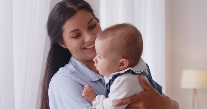 Loving young mother holding cute baby boy, embracing, stroking head with tenderness, spending time together at home, close up. Concept of happy motherhood and unconditional love between mom and kid