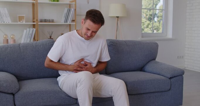 Sick young man pain in stomach or intestine sitting on sofa at home and clutching abdomen. Person suffering from abdominal pain and cramps in belly, sharp ache of gastroenterology diseases or bad diet