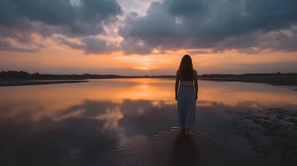 Poster Reflectie A woman walks into shallow water at sunset with dramatic clouds reflected in the calm surface  © Bussakon