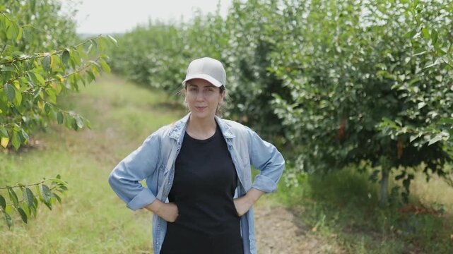A young white female farmer, wearing a baseball cap, is captured in an orchard. She is deeply involved in inspecting the plants, surrounded by the natural beauty of the farmland.