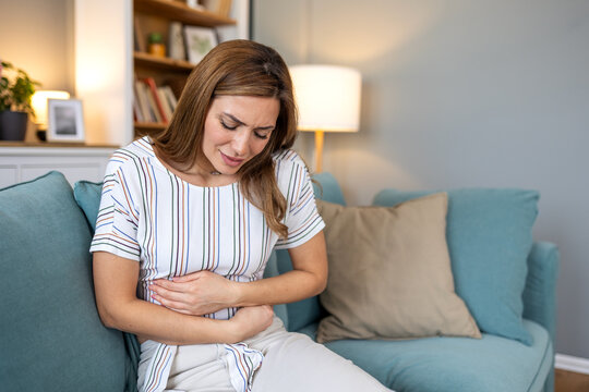 Stomach ache and digestion. Attractive woman sitting on sofa holding painful belly with hands, Healthcare And Medicine concept. Female person with abdominal problem feeling cramps and nausea at home.