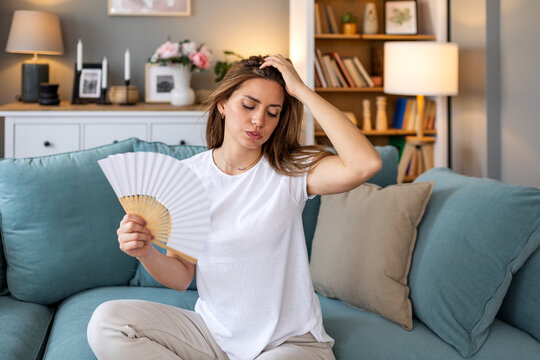 Menopause and hot flashes. Attractive woman sitting on sofa using hand fan to cool down, Healthcare And Medicine concept. Female person feeling hot and sweating from fever or heatwave in apartment.