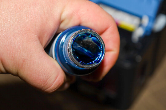 ​A male hand holds an open bottle of rich blue ink for refilling a printer, showcasing the paint's texture and sheen. This close-up shot emphasizes the consumable material's readiness for use in a con