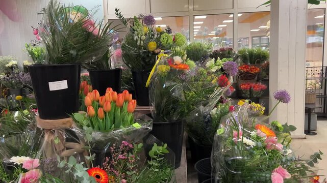 A variety of floral bouquets, including orange tulips and gerberas, are displayed in black buckets and plastic wraps at a busy flower market warehouse.