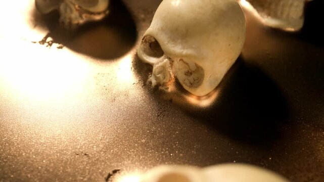 Dramatic panning shot moving slowly across a textured surface of shimmering golden paint that surrounds miniature human skulls creating a sea of gold effect where high contrast lighting