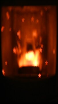 Vertical blurred video of a pellet stove with glowing fire burning inside, while soot forms a subtle star shape pattern on the glass. The soft focus enhances warmth, movement, and cozy indoor 