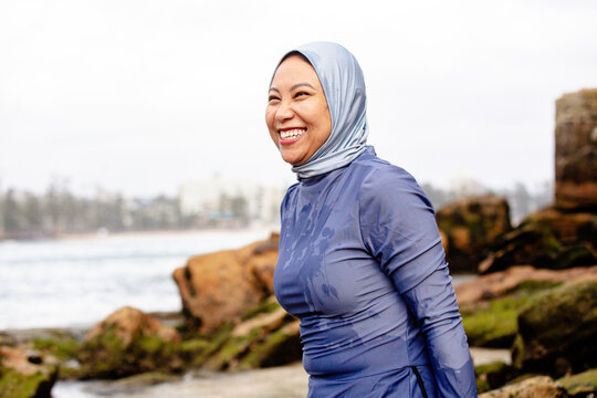 Happy Filipina woman wearing a burkini and light blue headscarf sitting on a rock on the beach