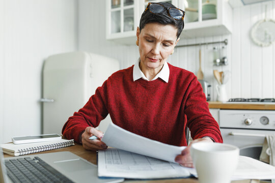 Indoor image of aged stylish accountant general making annual financial report sitting at kitchen table and working at home after coming back from office, reading and revising paper docs