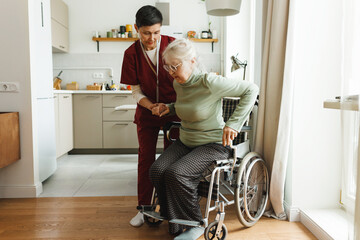 Indoor image of female nurse or physician in red uniform assisting her old patient at home helping...