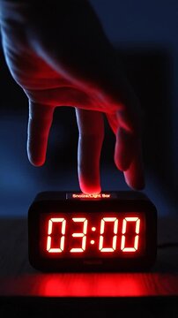 Close-up of a hand reaching to turn off a digital alarm clock showing 03:00 in the dark with a red glow