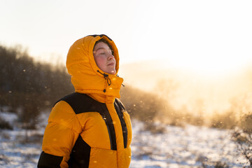 A traveler woman with closed eyes is standing outdoors breathing fresh air during a winter snowfall in the golden sunlight, meditating in the cold weather and feeling gratitude in nature. 