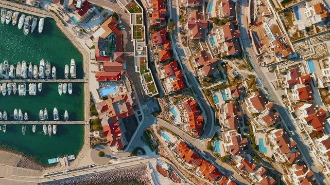 Aerial view of a Montenegro Adriatic resort marina with parallel piers, moored yachts, a curved breakwater, terracotta roofs, pools, winding roads, and late afternoon light.