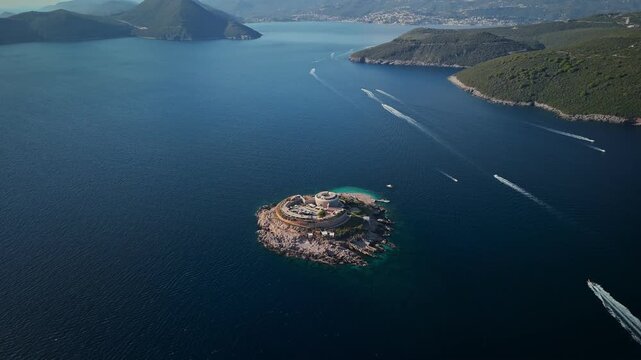 Aerial view of Mamula Fortress on Lastavica near the Bay of Kotor, Montenegro. Speedboats pass, wakes streak the water, wide view shows walls, rim, and coast.