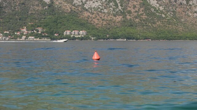 An orange conical buoy bobs as a white speedboat races by Montenegro's Adriatic shore, red roof buildings and pine on rocky hills in bright midday light.