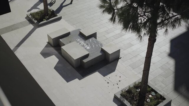 Elevated overhead view of a minimalist urban plaza in Montenegro with a geometric concrete fountain, raised tree planters, and a person walking a small dog.