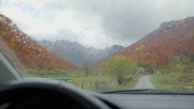 From the driver's seat, a narrow lane winds into a Balkan valley with orange and red autumn forest, a green gate, fence, rocky ridge, and low clouds in overcast light.