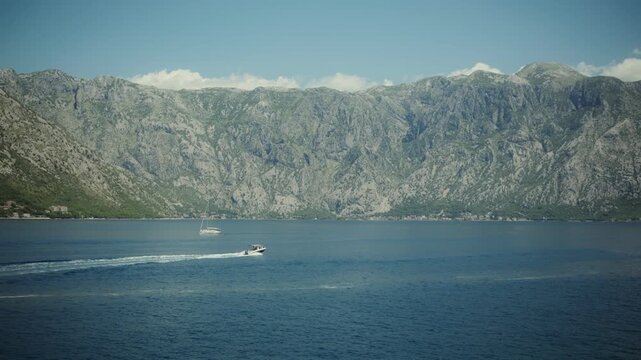 Daytime view of Bay of Kotor in Montenegro, motorboat speeds past with white wake, sailboat rests farther out, rugged karst cliffs and villages line the shore.