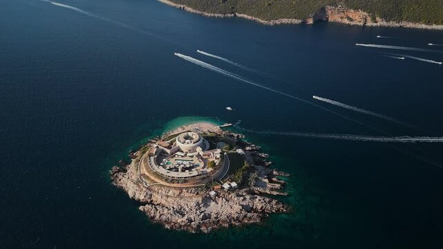Aerial view shows Mamula Lastavica off Montenegro, concentric terraces, round keep, pools and helipad, speedboats leave wakes, tender nears cove in warm late afternoon light.