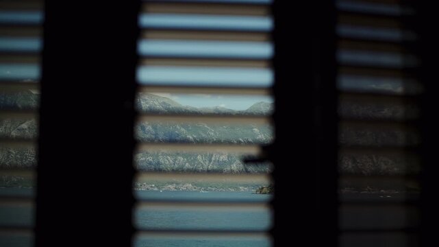 Calm Adriatic bay and waterfront village appear through Venetian blinds as the view shifts, revealing rocky karst mountains and limestone cliffs in Montenegro.