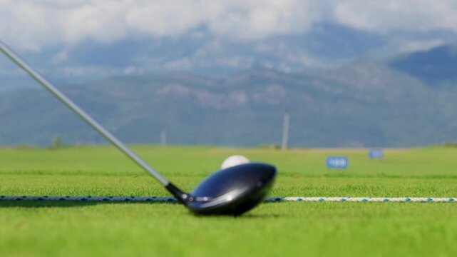 Low angle close up shows a driver lowering onto a teed ball at a neat range in Montenegro, 100 and 150 markers visible, daylight, shallow depth, mountain backdrop.