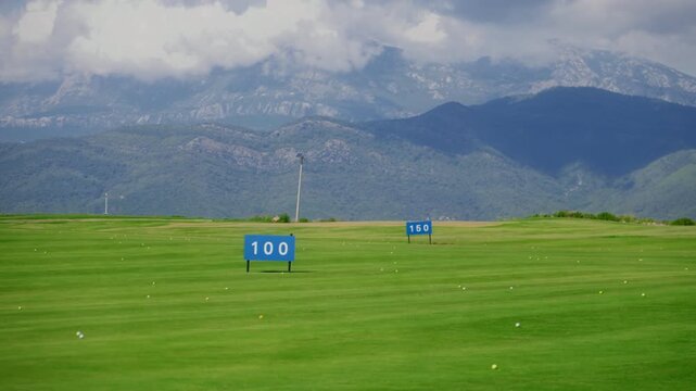 Wide daytime golf driving range with striped turf, many balls, 100 and 150 markers, rugged Montenegro mountains and clouds, broad frame, horizontal expanse.