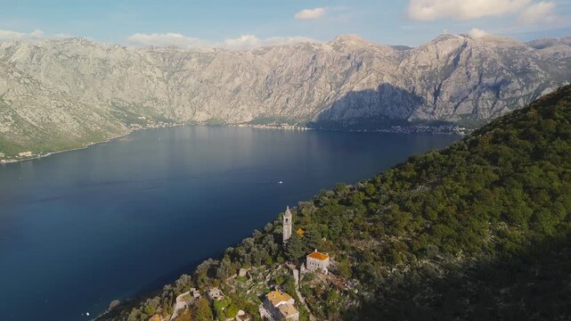 Aerial view of a stone village with orange roofs and bell tower on a steep hillside at Bay of Kotor, Montenegro. A motorboat moves as late sun casts a mountain shadow.