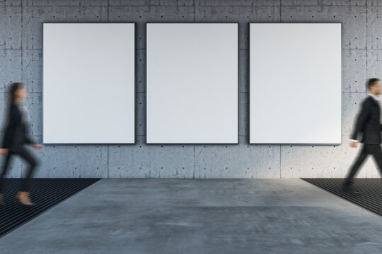 Three vertical blank posters on concrete wall in clean passway with blurred people walking by in a modern indoors area hall zone.""