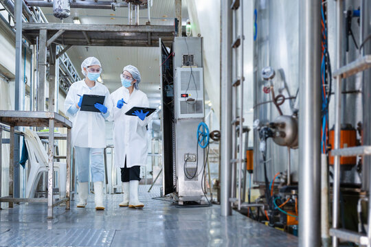Quality control team using digital tablet and clipboard for production line audit in modern plant, Female scientists wearing face masks and safety goggles working in high-standard processing facility
