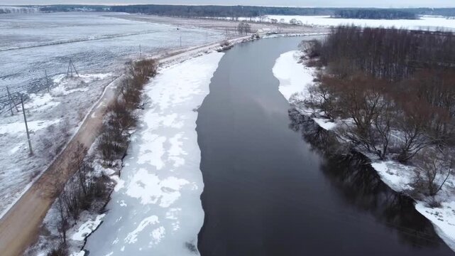View from above:  A river flows through a snowy landscape with patches of ice along the banks. Trees line the river and fields show a mix of snow and bare soil. The setting is quiet and still.