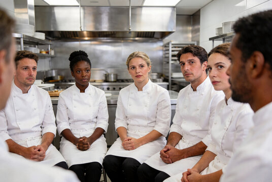 Diverse professional chefs in crisp white uniforms sitting in circular debriefing session within modern high-end kitchen with stainless steel elements. Candid atmosphere with expressive faces captured