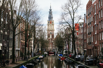 Amsterdam canal showing numerous boats moored along the waterfront, reflecting the classic gabled buildings and the iconic clock tower of the zuiderkerk against the cloudy sky