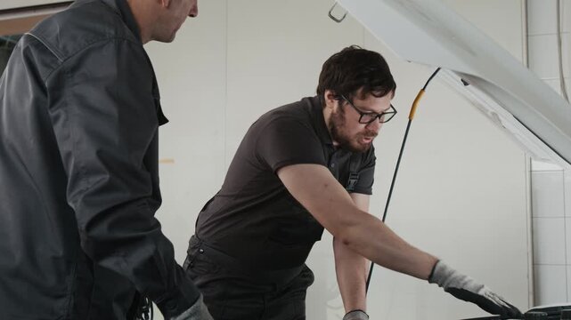 Medium side shot of two Caucasian male mechanics standing by car in garage, examining motor, discussing malfunction reason, young technician pointing at spark plugs to mature colleague
