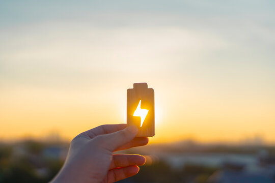 Renewable energy battery icon held against sunset sky. Concept of solar power charging, sustainable green technology, clean resource innovation, and future environmental solution for business.