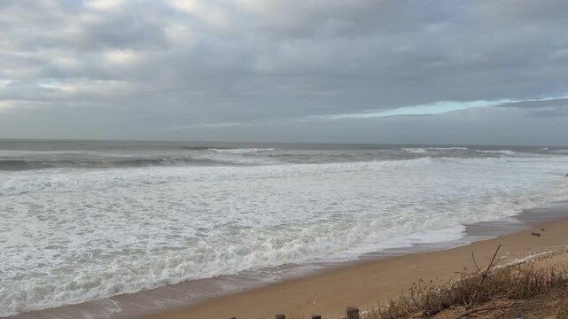 4K static shot of powerful Atlantic Ocean waves during high tide at Labenne beach, France. Massive sea foam and crashing water on the sandy shore under a cloudy sky