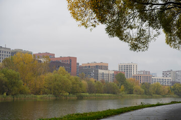 Obraz premium Golden colorful autumn in the city park with a modern residential area on the background. Footpath in a city park near the pond. Sustainable lifestyle in the town