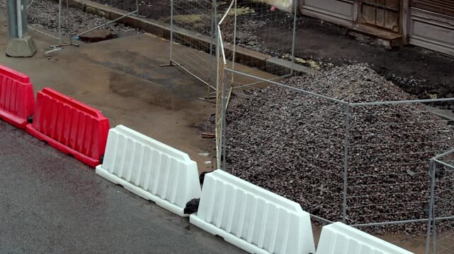 Mound of dark gravel is enclosed by metal mesh fencing, with white plastic barriers lining the adjacent pavement. The scene conveys a sense of temporary disruption in an urban environment, highlightin