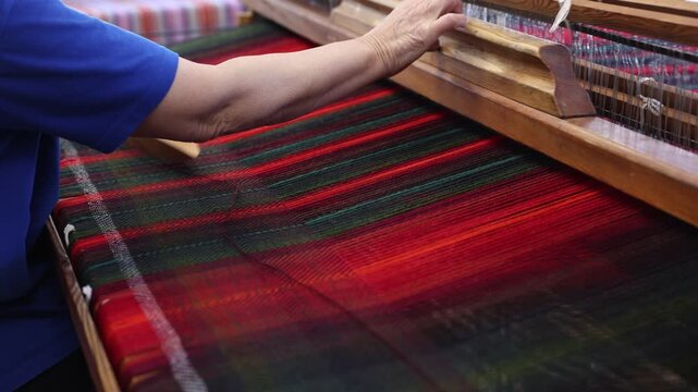 Close-up of a craftsperson weaving vibrant red and green tartan fabric on a traditional wooden loom. Hands guide the shuttle and threads in a textile workshop setting