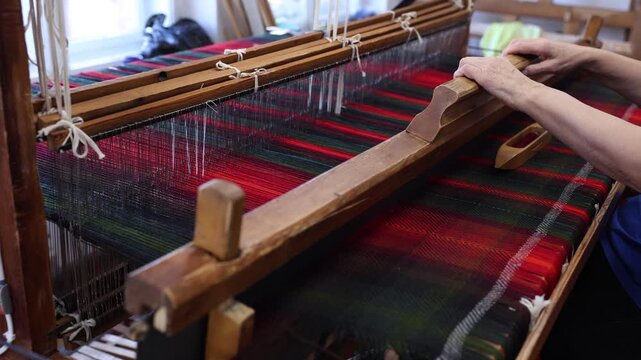 Close-up of a craftsperson weaving vibrant red and green tartan fabric on a traditional wooden loom. Hands guide the shuttle and threads in a textile workshop setting