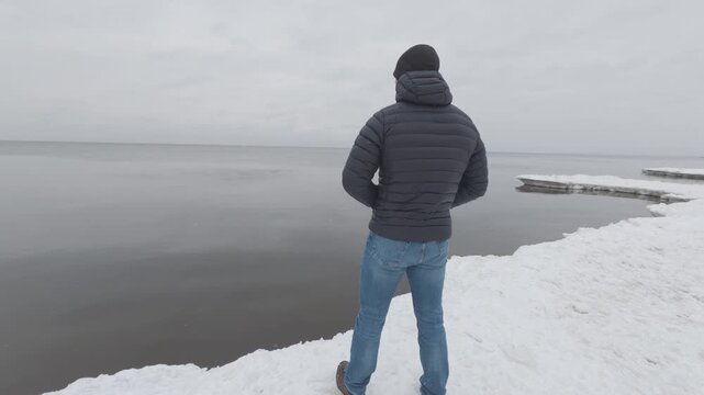 Man standing on snowy shore looking at sea, back view, winter landscape, calm cold weather, solitude and peaceful lifestyle concept